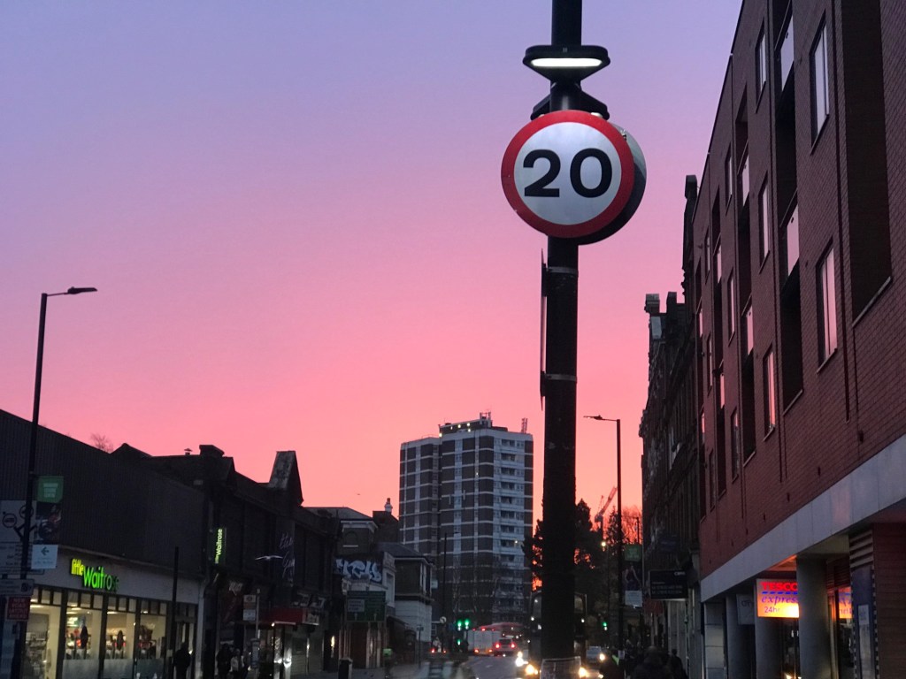 A street sign showing 20mph against a city backdrop and a pink sky